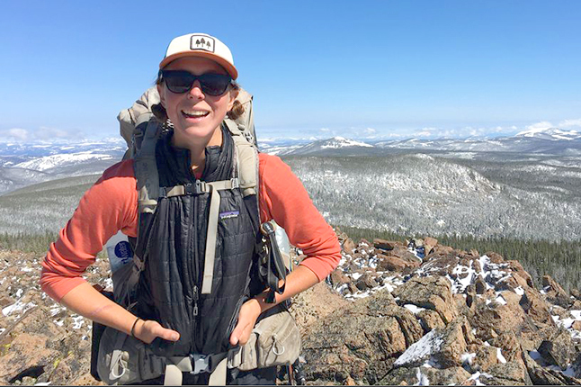 A woman smiles while standing on a ridge top with snowcapped mountains in the background