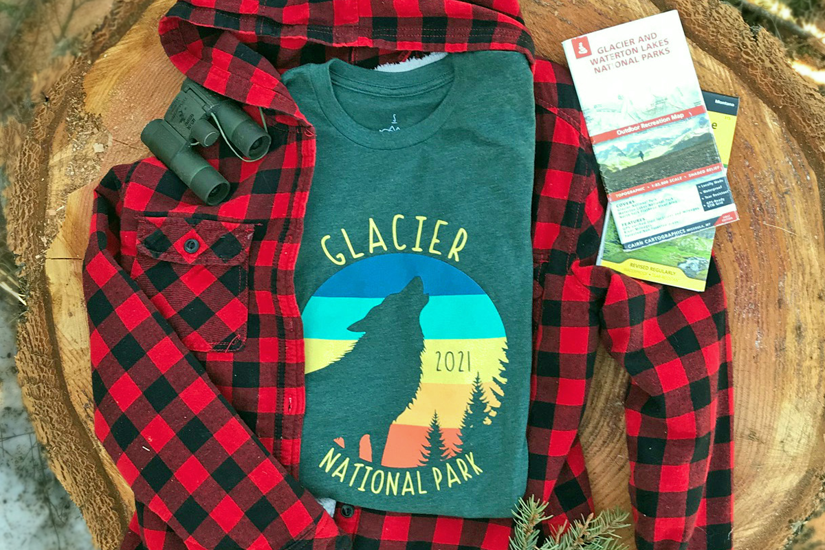 A forest green tee with wolf design and text that reads Glacier National Park 2021 lays on a tree stump outside next to red and black flannel, a pair of binoculars, and a variety of Glacier National Park maps