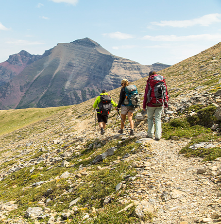 A group of three hikers traverse a trail with a prominent peak in the background