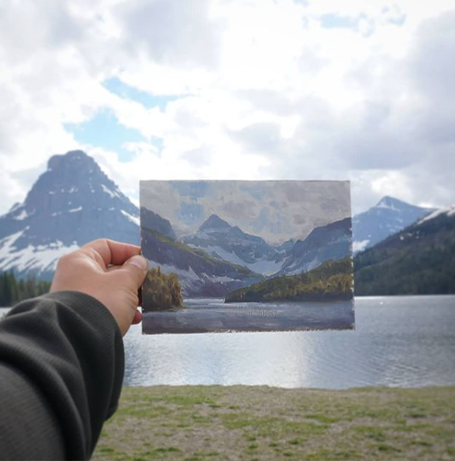 An artist holds out a plein air painting at the foot of a lake with a mountain landscape in the background