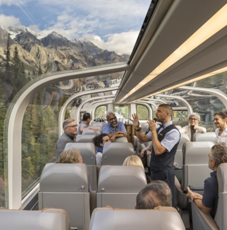 A group of train travelers gather around a host aboard the Rocky Mountaineer with a prominent mountain range in the background